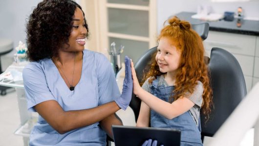 Medicine, pediatric dentistry and oral care concept. Happy female African dentist and curly red haired kid girl, having fun and giving high five, while holding tablet pc computer.